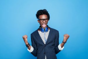 Studio portrait of successful afro american young man wearing striped top, navy blue jacket, nerd glasses, hat and headphone, laughing at camera with raised fists. Studio portrait, blue background.