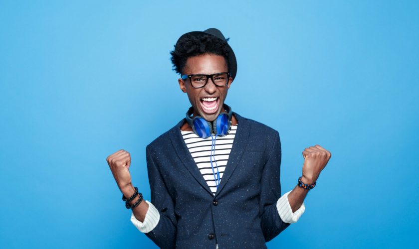 Studio portrait of successful afro american young man wearing striped top, navy blue jacket, nerd glasses, hat and headphone, laughing at camera with raised fists. Studio portrait, blue background.